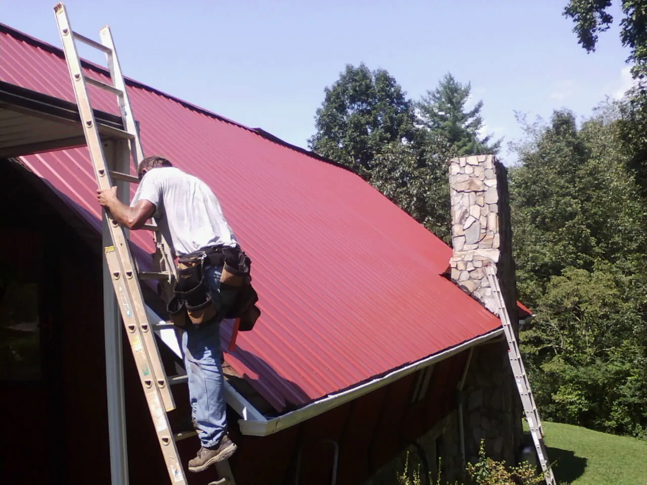 Roofer on a ladder beside a red metal roof and stone chimney — Soffit & Fascia Repair by Roofing Brothers in Sea Girt